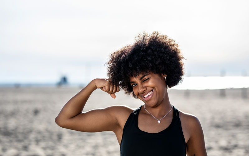 A woman of color with big curly hair makes a muscle pose and smiles while standing on a beach