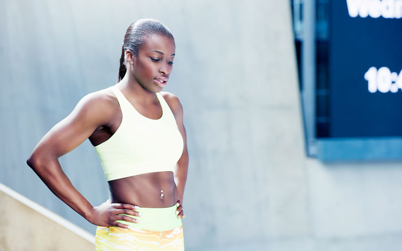 A woman of color in a sports bra and light yellow leggings looks pensive during an outdoor workout.